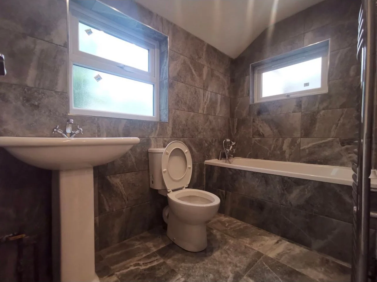 A fully tiled bathroom with a bath, pedestal basin, WC and a chrome heated towel rail on the wall